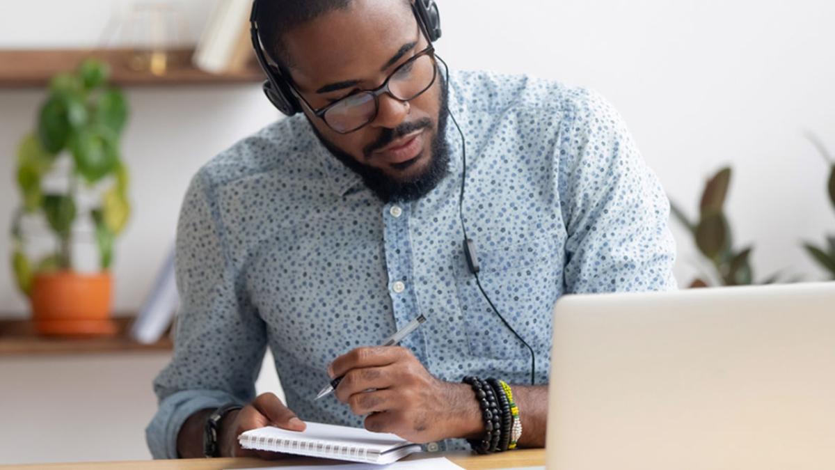 A man wearing headphones sitting in front of a computer and taking notes.