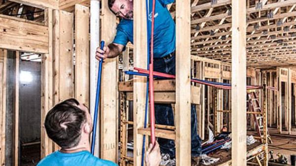 Two construction workers in blue shirts installing PEX piping in a wooden-framed building under construction.