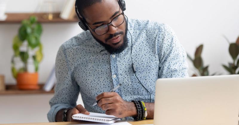 A man wearing headphones sitting in front of a computer and taking notes.